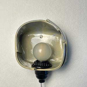 Puede incluir: Un aplique de pared con una pantalla de cer&aacute;mica en color verde salvia claro brillante, de forma rectangular redondeada y org&aacute;nica. Un casquillo negro sostiene una bombilla redonda de vidrio esmerilado y un cable transparente.