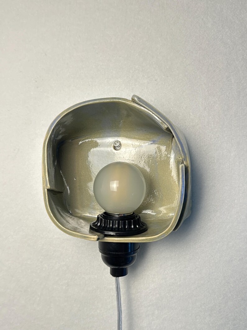 Puede incluir: Un aplique de pared con una pantalla de cer&aacute;mica en color verde salvia claro brillante, de forma rectangular redondeada y org&aacute;nica. Un casquillo negro sostiene una bombilla redonda de vidrio esmerilado y un cable transparente.