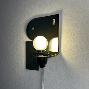 May include: A wall-mounted lamp featuring a glossy black ceramic backplate with a curved top edge and a rectangular base. A single round, frosted white light bulb is secured in a black socket, emitting a warm glow from the centre of the fixture.