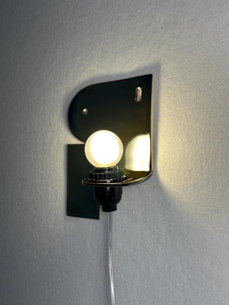 May include: A wall-mounted lamp featuring a glossy black ceramic backplate with a curved top edge and a rectangular base. A single round, frosted white light bulb is secured in a black socket, emitting a warm glow from the centre of the fixture.