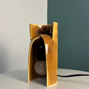 Puede incluir: Una l&aacute;mpara de mesa con una pantalla de cer&aacute;mica curva en color amarillo mostaza con un acabado brillante y moteado. La estructura tiene un dise&ntilde;o frontal abierto y arqueado que deja ver un casquillo negro y una bombilla blanca redonda.