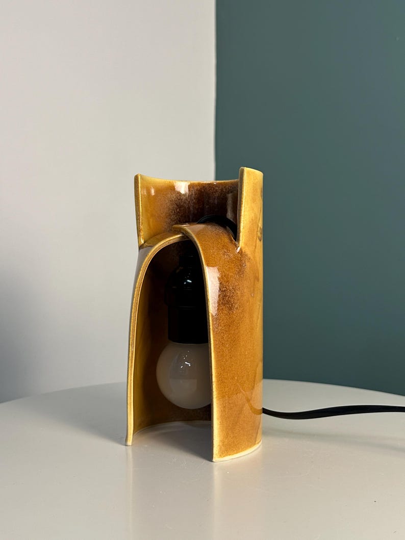 Puede incluir: Una l&aacute;mpara de mesa con una pantalla de cer&aacute;mica curva en color amarillo mostaza con un acabado brillante y moteado. La estructura tiene un dise&ntilde;o frontal abierto y arqueado que deja ver un casquillo negro y una bombilla blanca redonda.