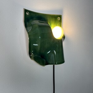 May include: A wall-mounted light fixture featuring a glossy, dark green, organic-shaped ceramic panel with two mounting holes at the top. A single, warm-glowing spherical light bulb is positioned within a cutout on the right side of the panel.