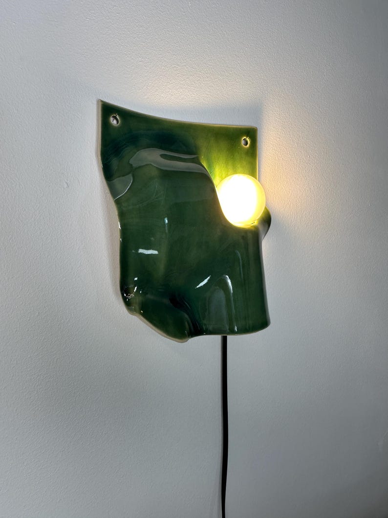 May include: A wall-mounted light fixture featuring a glossy, dark green, organic-shaped ceramic panel with two mounting holes at the top. A single, warm-glowing spherical light bulb is positioned within a cutout on the right side of the panel.