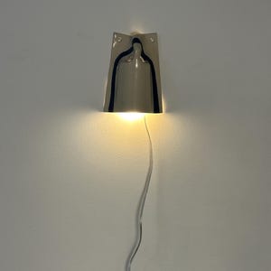 May include: A wall-mounted lamp with a polished silver-tone metallic finish. The fixture features a trapezoidal shape with a central cutout that emits a warm, downward-facing light. A thin, clear electrical cord hangs vertically from the base.