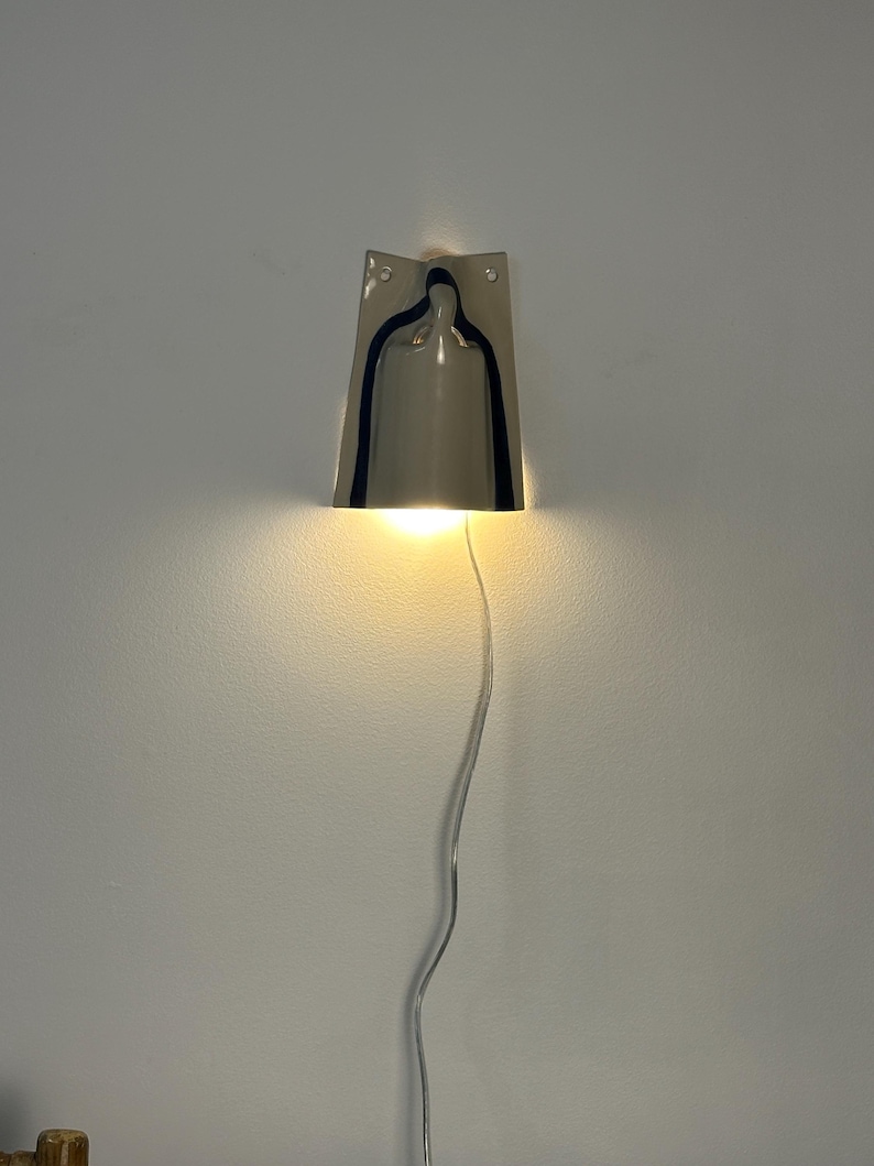 May include: A wall-mounted lamp with a polished silver-tone metallic finish. The fixture features a trapezoidal shape with a central cutout that emits a warm, downward-facing light. A thin, clear electrical cord hangs vertically from the base.