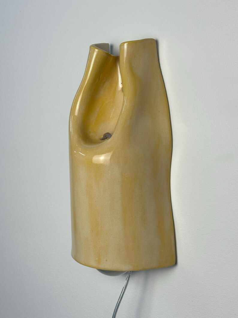 Pu&ograve; includere: Una lampada da parete in ceramica con una finitura lucida color giallo pallido. L'oggetto presenta una silhouette allungata, organica e curva con una sezione superiore scavata e una piccola apertura circolare. Un sottile cavo esce dalla base.