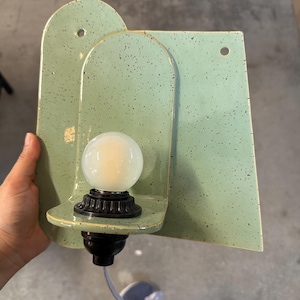 May include: A wall-mounted light fixture featuring a light green ceramic backplate with dark speckles. The fixture has an arched design with two circular mounting holes and a black socket holding a round, frosted white light bulb with a cord.