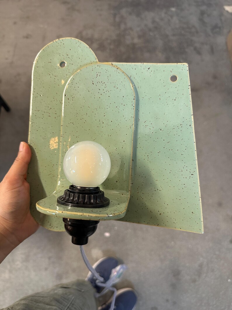 May include: A wall-mounted light fixture featuring a light green ceramic backplate with dark speckles. The fixture has an arched design with two circular mounting holes and a black socket holding a round, frosted white light bulb with a cord.