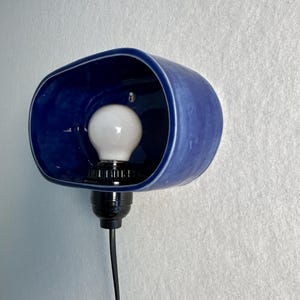 Może przedstawiać: Niebieski ceramiczny kinkiet z białą żar&oacute;wką. Kinkiet ma zaokrąglony, owalny kształt i jest zamontowany na białej ścianie. Żar&oacute;wka jest w czarnym gnieździe, a czarny przew&oacute;d zwisa w d&oacute;ł.