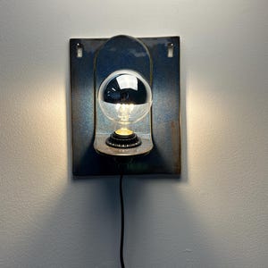 May include: A wall-mounted ceramic sconce featuring a rectangular blue-glazed backplate with a curved shelf. A clear light bulb with a reflective silver top cap is centered on the shelf, emitting a warm glow from the base and a black power cord.