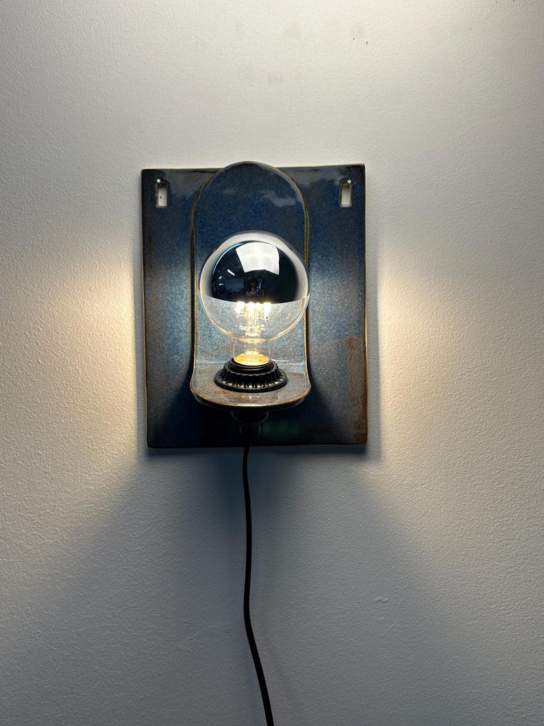 May include: A wall-mounted ceramic sconce featuring a rectangular blue-glazed backplate with a curved shelf. A clear light bulb with a reflective silver top cap is centered on the shelf, emitting a warm glow from the base and a black power cord.