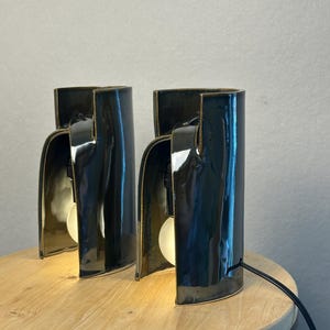 Puede incluir: Dos l&aacute;mparas de mesa de cer&aacute;mica de color gris oscuro brillante con una estructura curva semicil&iacute;ndrica. Cada l&aacute;mpara presenta un dise&ntilde;o frontal abierto que revela una bombilla blanca alojada dentro de la carcasa de cer&aacute;mica lisa y reflectante.
