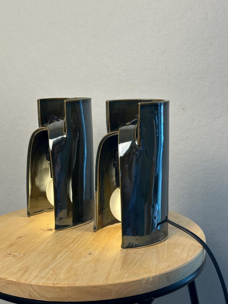 Puede incluir: Dos l&aacute;mparas de mesa de cer&aacute;mica de color gris oscuro brillante con una estructura curva semicil&iacute;ndrica. Cada l&aacute;mpara presenta un dise&ntilde;o frontal abierto que revela una bombilla blanca alojada dentro de la carcasa de cer&aacute;mica lisa y reflectante.