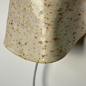 May include: A wall-mounted ceramic light fitting with a glossy, off-white glaze featuring brown speckles. The lamp has a curved, organic shape and emits a warm, glowing light from the top opening, with a thin power cord extending downwards.