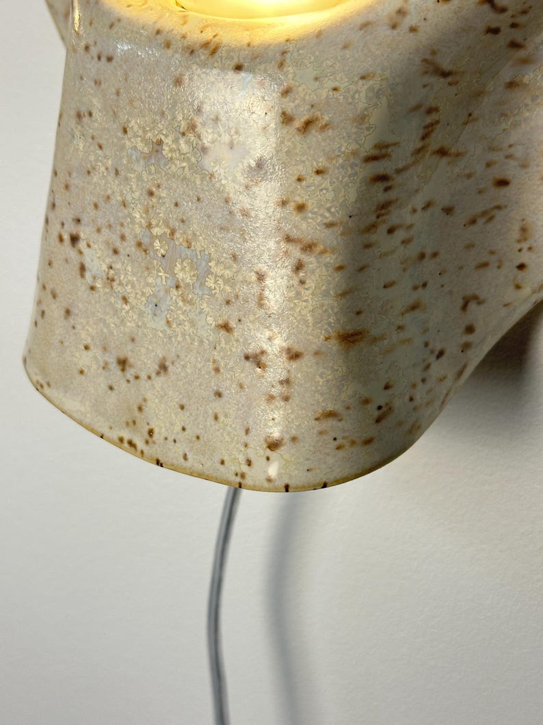 May include: A wall-mounted ceramic light fitting with a glossy, off-white glaze featuring brown speckles. The lamp has a curved, organic shape and emits a warm, glowing light from the top opening, with a thin power cord extending downwards.