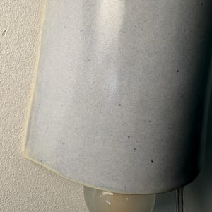 K&ouml;nnte beinhalten: Eine Wandleuchte mit einem geschwungenen, hellblauen Keramikschirm mit gesprenkelter Textur. Der Schirm hat eine offene Unterseite, die eine kleine, runde Gl&uuml;hbirne aus Milchglas freigibt. Ein transparentes Stromkabel h&auml;ngt von der Leuchte.