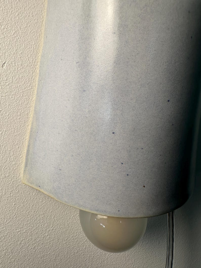 K&ouml;nnte beinhalten: Eine Wandleuchte mit einem geschwungenen, hellblauen Keramikschirm mit gesprenkelter Textur. Der Schirm hat eine offene Unterseite, die eine kleine, runde Gl&uuml;hbirne aus Milchglas freigibt. Ein transparentes Stromkabel h&auml;ngt von der Leuchte.