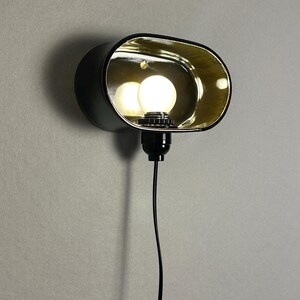 K&ouml;nnte beinhalten: Schwarze Wandleuchte mit goldfarbenem Innenraum und einer runden Gl&uuml;hbirne. Die Leuchte hat eine ovale Form und ein schwarzes Netzkabel. Das Licht ist eingeschaltet und beleuchtet den goldenen Innenraum.