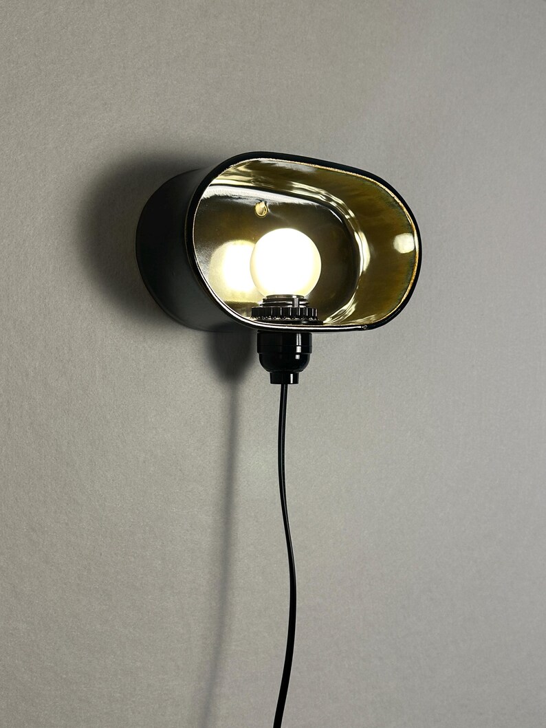 K&ouml;nnte beinhalten: Schwarze Wandleuchte mit goldfarbenem Innenraum und einer runden Gl&uuml;hbirne. Die Leuchte hat eine ovale Form und ein schwarzes Netzkabel. Das Licht ist eingeschaltet und beleuchtet den goldenen Innenraum.
