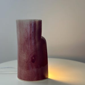 May include: A cylindrical ceramic vase with a glossy, deep magenta and plum-colored glaze. The vase features an asymmetrical, stepped silhouette with a taller main section and a shorter, rounded side extension, emitting a warm glow from below.
