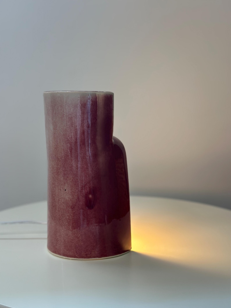 May include: A cylindrical ceramic vase with a glossy, deep magenta and plum-colored glaze. The vase features an asymmetrical, stepped silhouette with a taller main section and a shorter, rounded side extension, emitting a warm glow from below.