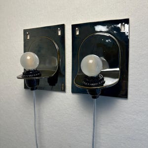May include: Two wall-mounted sconces with a dark blue-green finish. Each sconce features a square backplate, a curved shelf, and a round, frosted light bulb. The sconces are connected to a wall outlet via a gray cord.