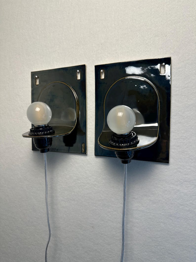 May include: Two wall-mounted sconces with a dark blue-green finish. Each sconce features a square backplate, a curved shelf, and a round, frosted light bulb. The sconces are connected to a wall outlet via a gray cord.