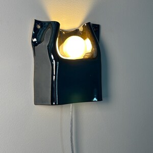May include: A wall-mounted sconce featuring a glossy, dark-colored ceramic body with an irregular, wavy top edge. A single round, warm-toned light bulb is recessed within the center, casting a soft glow upward. A thin, clear power cord hangs below.
