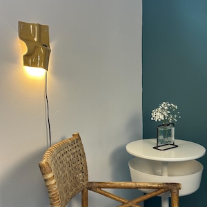 K&ouml;nnte beinhalten: Eine senfgelbe Wandleuchte aus Keramik mit gl&auml;nzender Oberfl&auml;che und warm leuchtender Gl&uuml;hbirne, die eine geschwungene, organische Form aufweist und mit einem schwarzen Stromkabel vertikal &uuml;ber einem Rattanstuhl an der Wand befestigt ist.