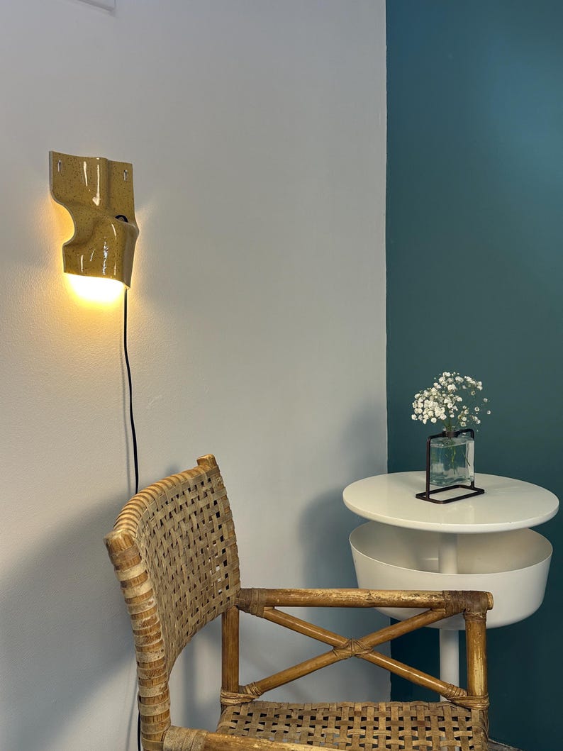 K&ouml;nnte beinhalten: Eine senfgelbe Wandleuchte aus Keramik mit gl&auml;nzender Oberfl&auml;che und warm leuchtender Gl&uuml;hbirne, die eine geschwungene, organische Form aufweist und mit einem schwarzen Stromkabel vertikal &uuml;ber einem Rattanstuhl an der Wand befestigt ist.