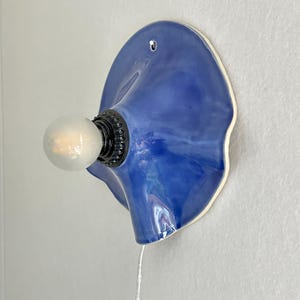 May include: A blue ceramic wall sconce with a bulb. The sconce has a unique, organic shape with a glossy finish. The bulb is round and emits a soft glow. The sconce is mounted on a white wall.