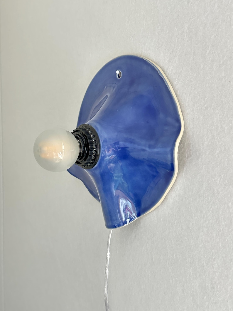 May include: A blue ceramic wall sconce with a bulb. The sconce has a unique, organic shape with a glossy finish. The bulb is round and emits a soft glow. The sconce is mounted on a white wall.