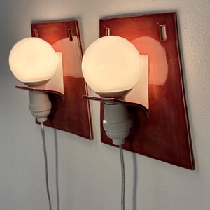 Puede incluir: Un par de apliques de pared con placas traseras de cer&aacute;mica de color rojo oscuro brillante y forma trapezoidal. Cada l&aacute;mpara incluye un casquillo blanco, un soporte curvo y una bombilla redonda de cristal esmerilado con cable el&eacute;ctrico.