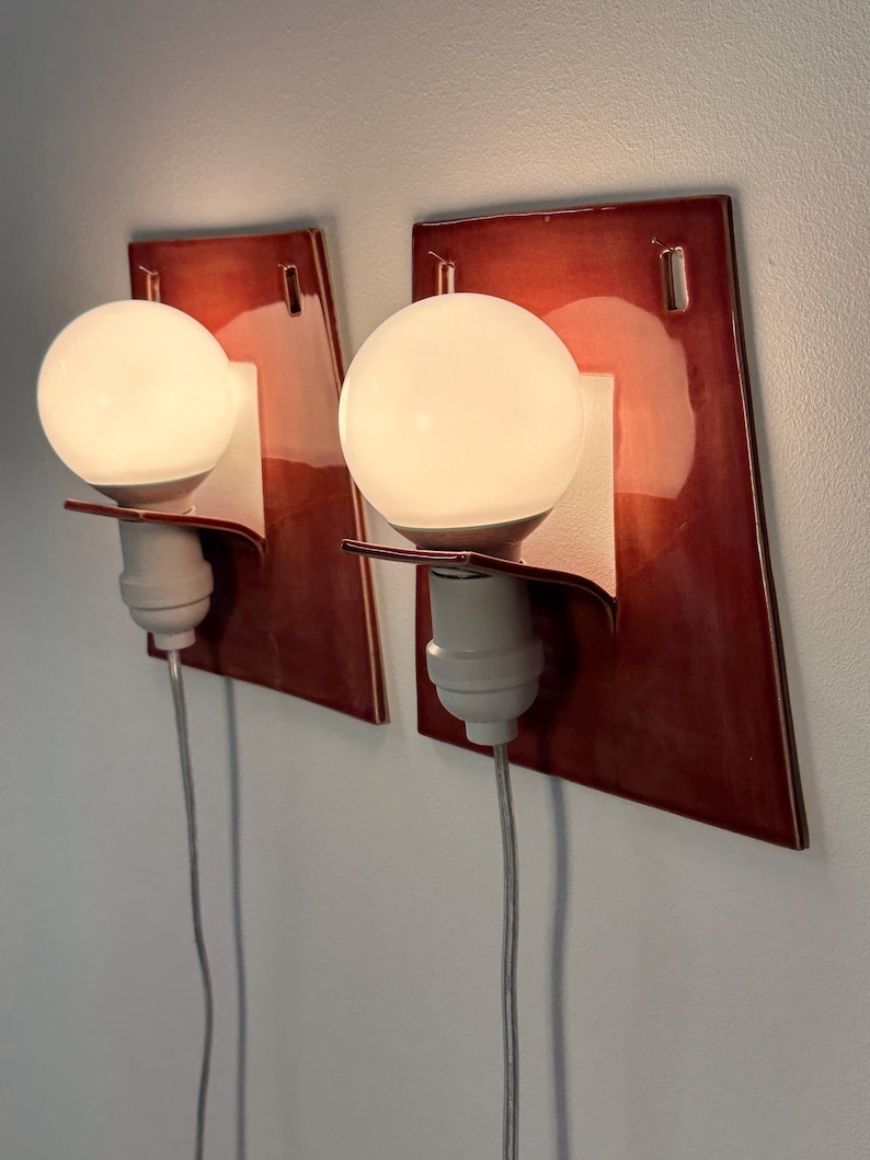 Puede incluir: Un par de apliques de pared con placas traseras de cer&aacute;mica de color rojo oscuro brillante y forma trapezoidal. Cada l&aacute;mpara incluye un casquillo blanco, un soporte curvo y una bombilla redonda de cristal esmerilado con cable el&eacute;ctrico.