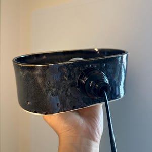 May include: A black ceramic light fitting with a glossy finish. The fitting has an oval shape and a black cord. The interior is white. The fitting is held by a hand.
