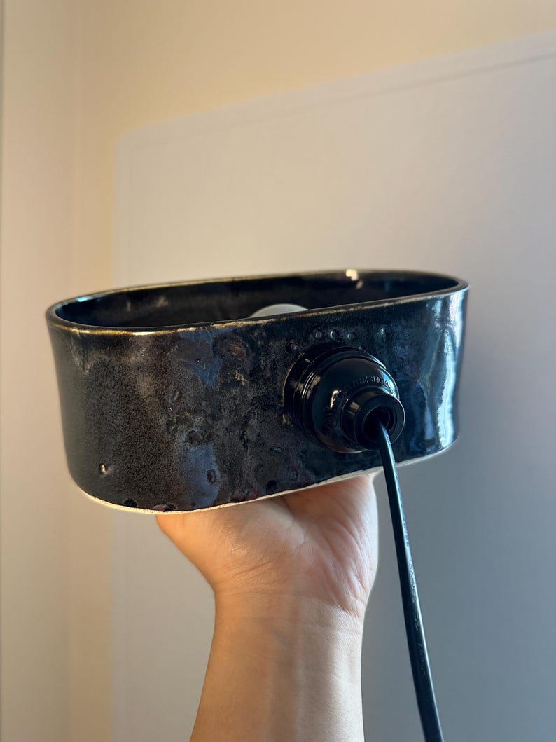 May include: A black ceramic light fitting with a glossy finish. The fitting has an oval shape and a black cord. The interior is white. The fitting is held by a hand.