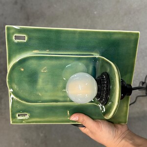 May include: A rectangular green ceramic wall sconce with a glossy finish, featuring a central U-shaped indentation and two small rectangular cutouts on the left side. A black light socket holds a round, frosted white light bulb in the center.