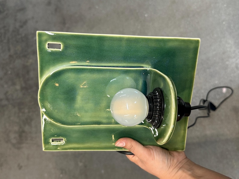 May include: A rectangular green ceramic wall sconce with a glossy finish, featuring a central U-shaped indentation and two small rectangular cutouts on the left side. A black light socket holds a round, frosted white light bulb in the center.