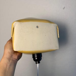 May include: A yellow ceramic wall-mounted lamp with a rectangular shape and rounded edges. The front face is flat and off-white with a small circular hole near the top center. A black light socket extends from the bottom with a clear power cord.