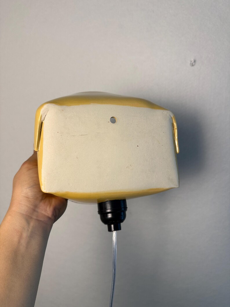 May include: A yellow ceramic wall-mounted lamp with a rectangular shape and rounded edges. The front face is flat and off-white with a small circular hole near the top center. A black light socket extends from the bottom with a clear power cord.