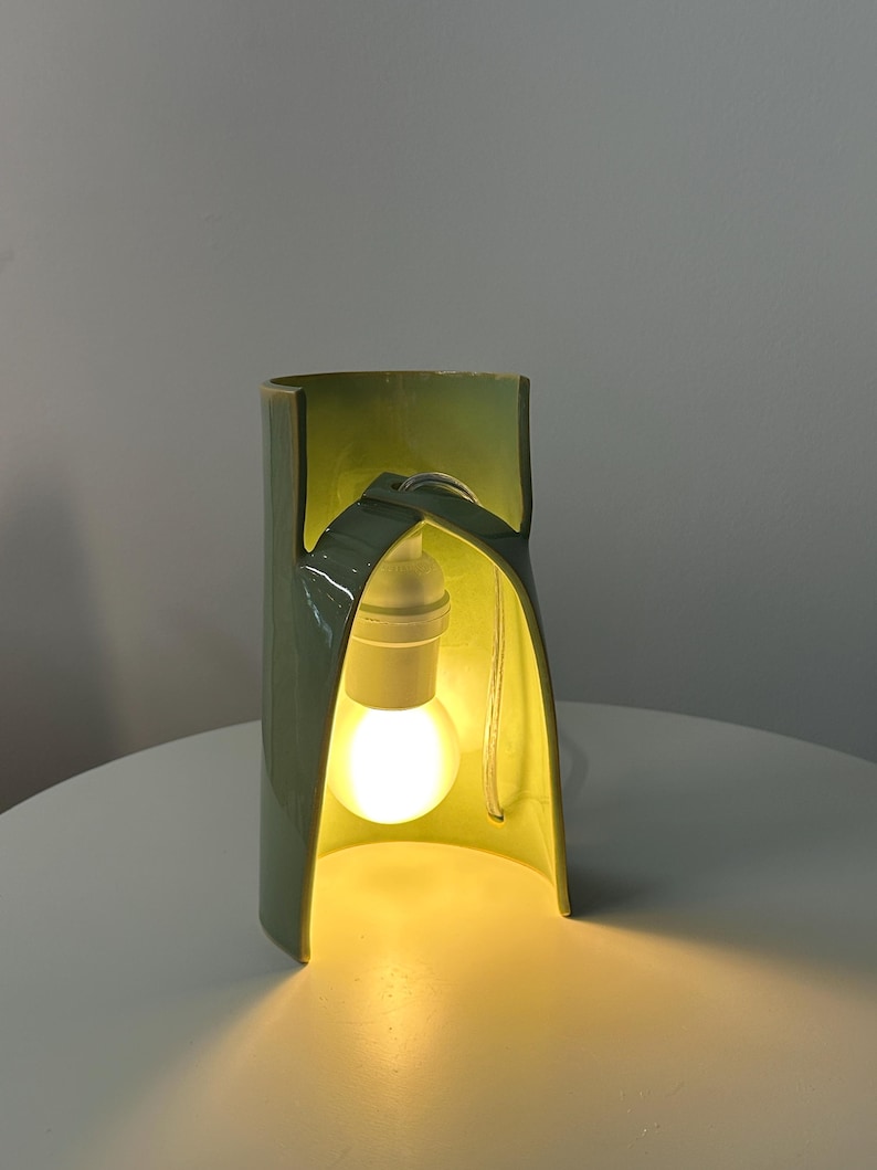 May include: A sage green ceramic table lamp with a curved, open-arched structure. The lamp features a white socket and a glowing light bulb suspended within the center of the arched ceramic frame, casting a warm light onto the surface below.