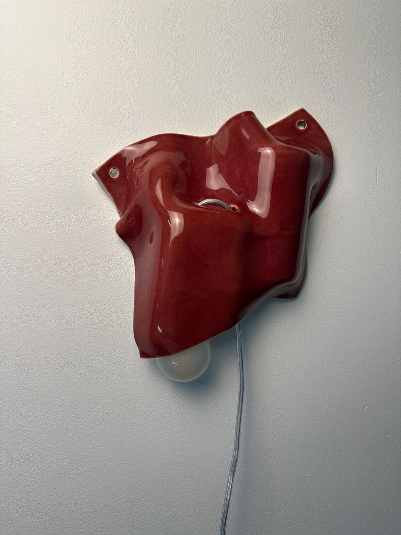 Puede incluir: Una l&aacute;mpara de pared con una pantalla de cer&aacute;mica de color rojo intenso brillante con una forma irregular, org&aacute;nica y plegada. El aplique incluye una peque&ntilde;a bombilla blanca redonda que sobresale por debajo y un cable el&eacute;ctrico fino.
