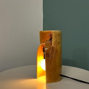 Puede incluir: Una l&aacute;mpara de mesa cil&iacute;ndrica de cer&aacute;mica con un esmalte brillante moteado en tonos &aacute;mbar y marr&oacute;n dorado. La l&aacute;mpara presenta una abertura vertical que revela una bombilla encendida en su interior y un cable de alimentaci&oacute;n negro.