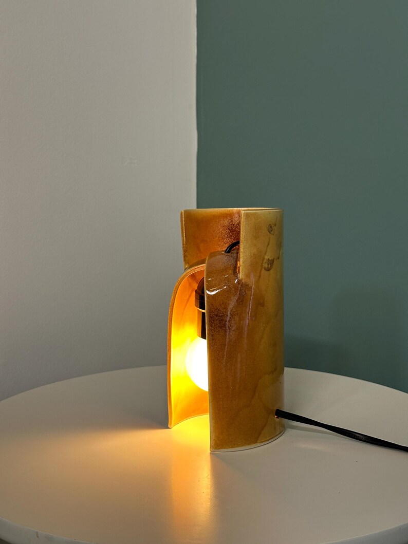 Puede incluir: Una l&aacute;mpara de mesa cil&iacute;ndrica de cer&aacute;mica con un esmalte brillante moteado en tonos &aacute;mbar y marr&oacute;n dorado. La l&aacute;mpara presenta una abertura vertical que revela una bombilla encendida en su interior y un cable de alimentaci&oacute;n negro.