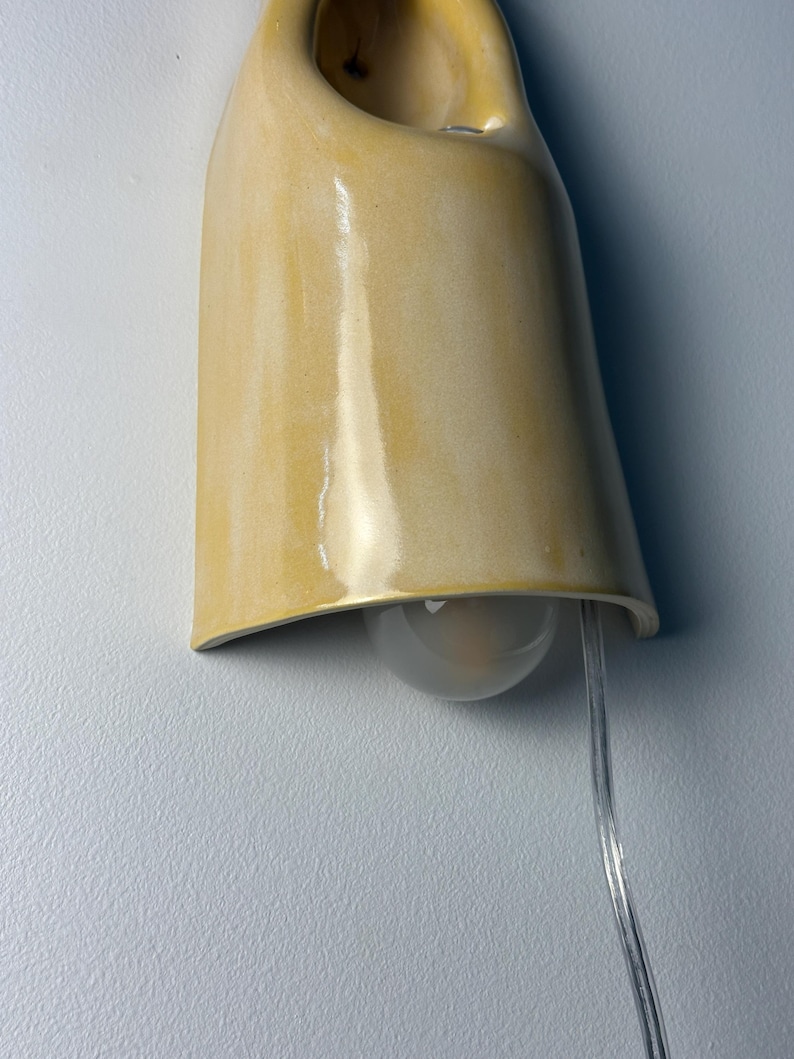 Pu&ograve; includere: Una lampada da parete con un paralume in ceramica gialla lucida e curva con una finitura liscia. L'apparecchio presenta un design aperto nella parte superiore e un cavo elettrico trasparente che fuoriesce dal basso, con una lampadina satinata.