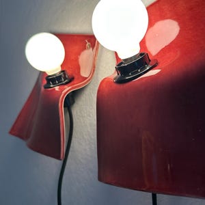 Puede incluir: Dos l&aacute;mparas de pared con pantallas de cer&aacute;mica curvadas en color rojo intenso y brillante. Cada l&aacute;mpara tiene un casquillo negro que sostiene una bombilla blanca redonda e iluminada. Montadas en la pared, incluyen cables negros.