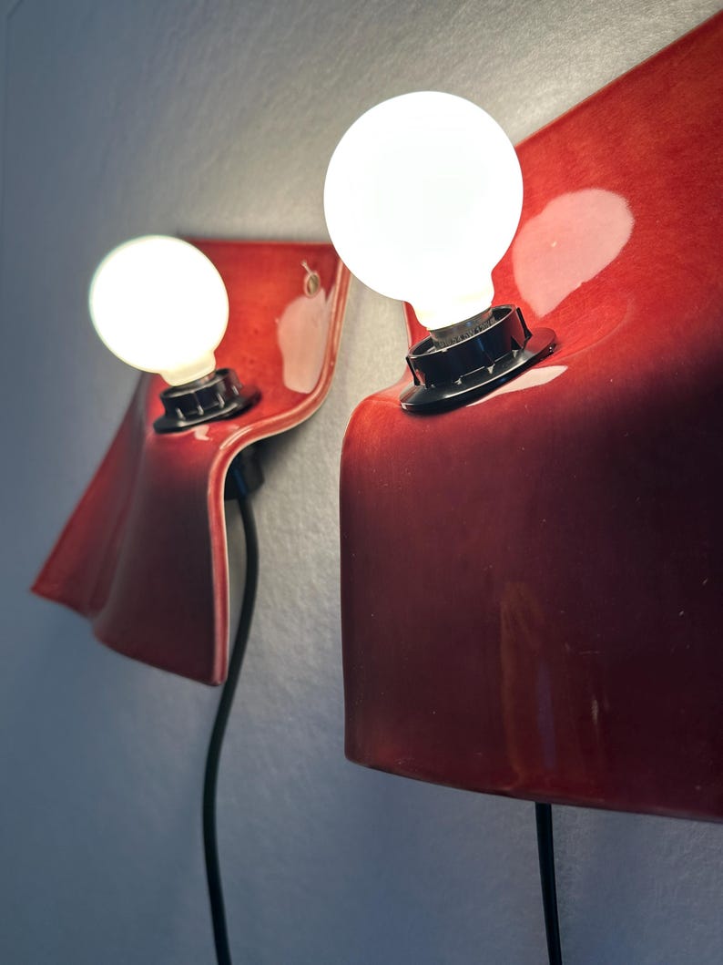 Puede incluir: Dos l&aacute;mparas de pared con pantallas de cer&aacute;mica curvadas en color rojo intenso y brillante. Cada l&aacute;mpara tiene un casquillo negro que sostiene una bombilla blanca redonda e iluminada. Montadas en la pared, incluyen cables negros.