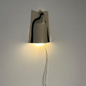 May include: A wall-mounted light fixture featuring a glossy, light gray ceramic shade with a curved, bell-like silhouette. The shade is accented by dark, vertical black stripes along the sides and emits a warm, downward-facing glow.