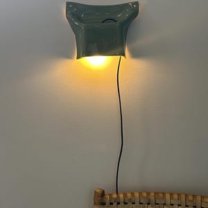 May include: A sage green ceramic wall sconce with a flared, sculptural shape. The fixture emits a warm, downward-facing glow and features a thin black power cord extending from the base. The light is mounted on a plain wall above a woven rattan surface.